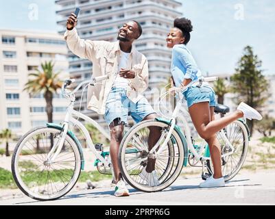 Selfie, bicicleta y cita con una pareja en bicicleta en un paseo marítimo durante el verano con amor, romance y afecto. Bicicleta, fotografía y diversión con una mujer negra y un hombre fuera y una ciudad en el fondo Foto de stock