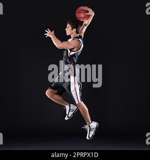 SLAM Dunk. Tiro de perfil completo de un joven jugador de baloncesto en acción contra un fondo negro. Foto de stock