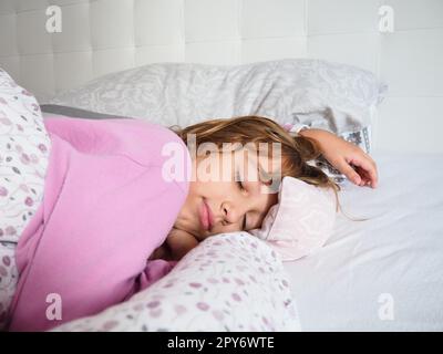 Una hermosa chica caucásica de 8 años de edad con el pelo rubio, vestida con pijama rosa, duerme en una cama con una manta esponjosa, abrazando una almohada. La luz suave del sol de la mañana fluye a través de la ventana. Foto de stock