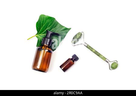Botellas cosméticas marrones, hoja verde y rodillo de cara de jade sobre fondo blanco. Vista superior, colocación plana. Foto de stock