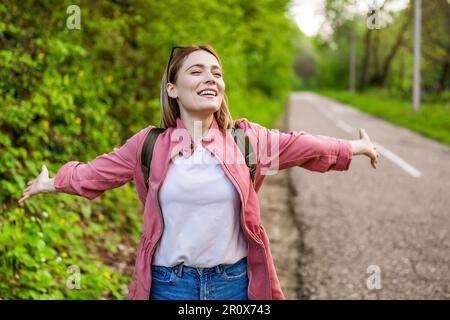 La mujer feliz está lista para caminar en el bosque. Ella está de pie junto a la carretera con los brazos extendidos. Foto de stock