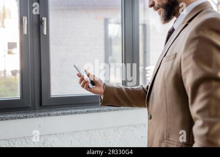 vista parcial del hombre de negocios barbudo en blazer elegante beige que sostiene el teléfono móvil con la pantalla en blanco mientras que está de pie cerca de grandes ventanas en contemporáneo o Foto de stock