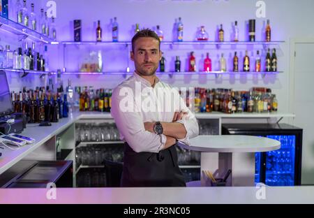 Barkeeper masculino positivo en uniforme y con las manos cruzadas sonriendo y mirando a cámara mientras se encuentra de pie en el bar moderno con varias marcas de licor bot Foto de stock