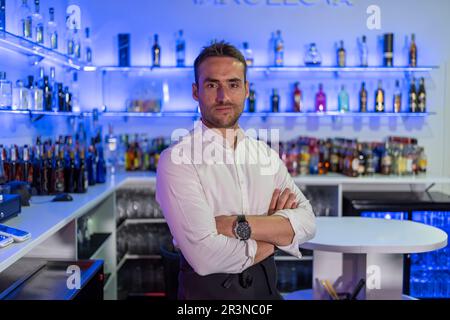 Barkeeper masculino positivo en uniforme y con las manos cruzadas sonriendo y mirando a cámara mientras se encuentra de pie en el bar moderno con varias marcas de licor bot Foto de stock