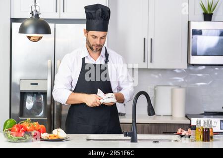 Hombre en delantal y sombrero de chef cocinando comida en cocina. Hombre  guapo que cocina la comida sana en la cocina. Chico cocinando comida de  cena en la cocina. Menú de inicio