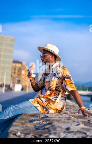 Retrato de un hombre étnico negro Disfrute de las vacaciones de verano en la playa comiendo un helado, sentado junto al río en un tiempo de vacaciones Foto de stock