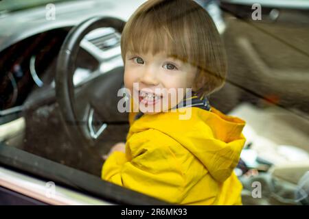 Adorable niño pequeño jugando en el asiento del conductor. Lindo pequeño hijo sentado en el coche del padre. Niño pequeño divirtiéndose en un vehículo. Foto de stock