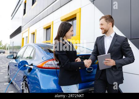 Mujer sonriente de pie en el coche y apuntando a la tableta del vendedor del coche. Ella es elegida el coche adecuado para ella que vio en línea. Una mujer concluye un acuerdo a. Foto de stock