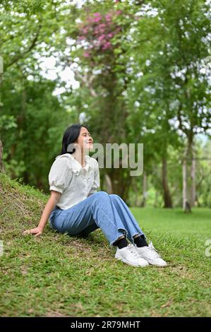Una mujer asiática joven atractiva y relajada en ropa casual se sienta en la hierba debajo de un árbol en un parque, respirando aire fresco, refrescándose, y f Foto de stock
