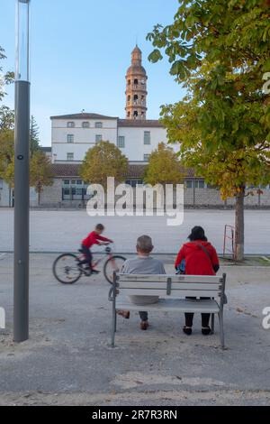 Cahors, FRANCIA - 29 de octubre de 2022: Dos personas sentadas en un banco y un niño montando una bicicleta al final del día en la plaza principal de Cahors en el m Foto de stock