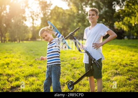 Dos niños se están divirtiendo con el monopatín y scooter en el parque. Niños juguetones en el parque, infancia feliz. Foto de stock