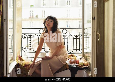 En una habitación de hotel de París, una mujer asiática de mediana edad glamorosa se sienta en un balcón Julieta con ventanas francesas abiertas con vistas a Grenelle en París, Francia Foto de stock