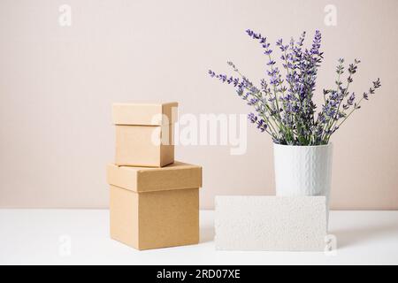Ramo de lavanda en un jarrón, maqueta de tarjeta de papel casera, cajas de regalo en una mesa blanca contra una pared de color crema. Foto de stock