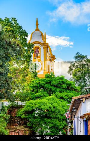 Torre de iglesia barroca que se eleva a través de la vegetación y los edificios en la ciudad de Tiradentes en el estado de Minas Gerais, Brasil Foto de stock