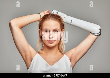 Retrato de estudio de primer plano de hermosa joven rubia con discapacidad que lleva brazo protésico biónico sensorial. Mujer bonita en vestido blanco posando con Foto de stock