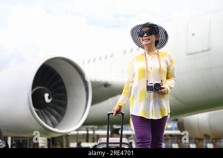 Mujer feliz de los sesenta en ropa de verano y gafas de sol caminando cerca del aeropuerto con un concepto de suitcase.Travel y vacaciones de verano Foto de stock