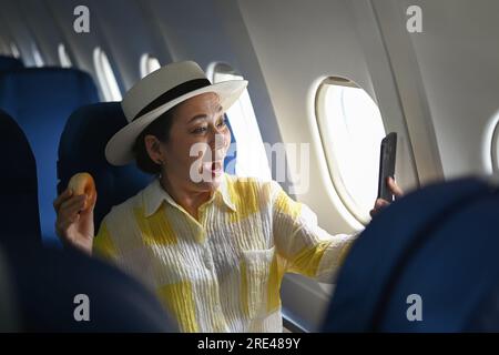 Turista mayor alegre de la mujer que toma selfie con el teléfono móvil mientras que se sienta en cabina del avión cerca de la ventana. Foto de stock