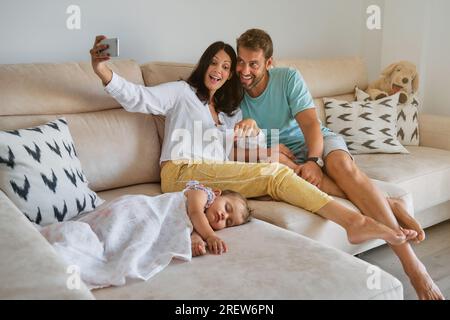 Mujer y hombre descalzos alegres que se divierten mientras toman selfie en el teléfono móvil y se sientan en el sofá cerca de la niña que siesta cubierta con manta en la vida Foto de stock