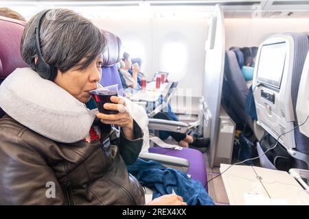 Mujer asiática bebiendo vino tinto mientras disfruta del entretenimiento a bordo durante el viaje en avión Foto de stock