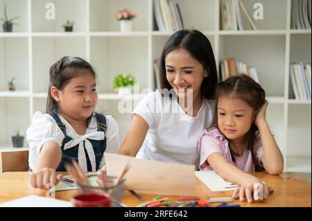 Una niñera asiática joven feliz y amable o maestra privada está enseñando arte a los niños pequeños, disfrutando del dibujo y la coloración juntos. Foto de stock