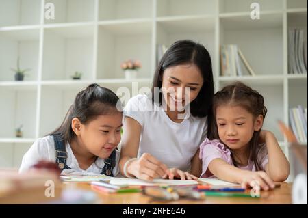 Una niñera asiática joven feliz y amable o maestra privada está enseñando arte a los niños pequeños, disfrutando del dibujo y la coloración juntos. niños, kin Foto de stock