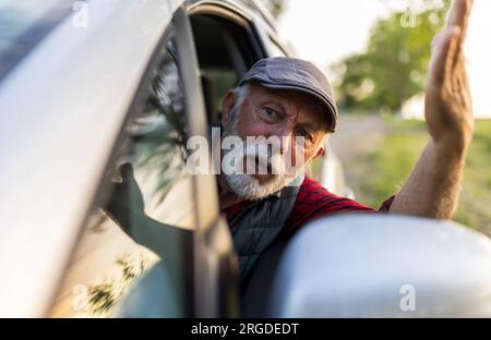 Hombre mayor que conduce el coche, mirando a través de la ventana lateral y discutiendo con alguien Foto de stock
