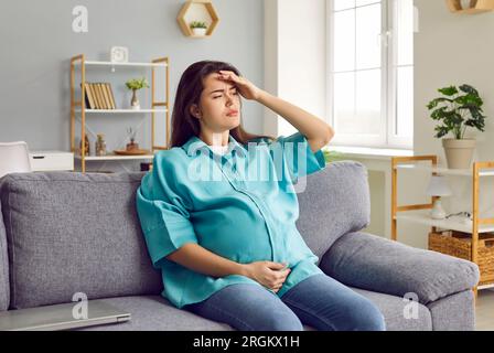 Mujer embarazada joven sentada en el sofá, teniendo un dolor de cabeza, sintiéndose un poco enferma y cansada Foto de stock