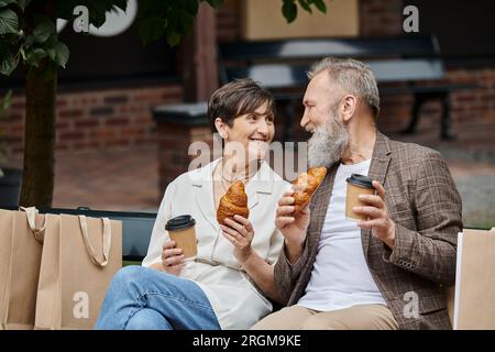 feliz pareja de ancianos sosteniendo croissants y café para ir, tazas de papel, bolsas de compras, outlet, urbano Foto de stock