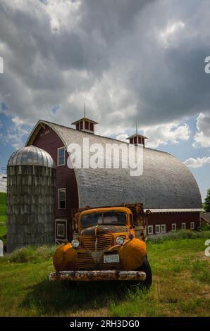 Un viejo camión naranja oxidado y un gran granero rojo (construido en 1919) con techo de cedro, dos cúpulas y un pequeño silo. Colfax, Washington, EE.UU. Foto de stock