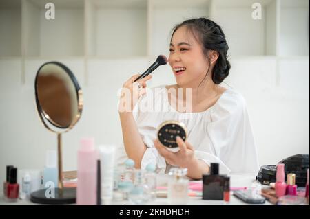 Una hermosa y alegre influencer asiática de belleza aplica rubor a sus mejillas mientras se hace el maquillaje en su habitación. Foto de stock