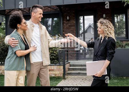 pareja interracial encantada tomando la llave de la nueva casa de campo del corredor de bienes raíces en la calle urbana Foto de stock