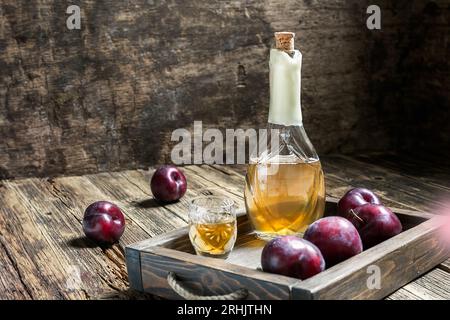 Brandy tradicional de ciruela de los Balcanes - rakija o rakia slivovica en la botella, una copa de vino con sljivovica y ciruelas frescas sobre el fondo de madera oscura Foto de stock