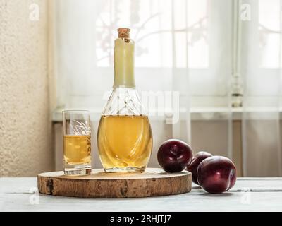 Brandy de ciruela balcánica - rakija o rakia slivovica en la botella, una copa de vino con sljivovica y ciruelas frescas en el fondo claro cerca de la ventana. Foto de stock