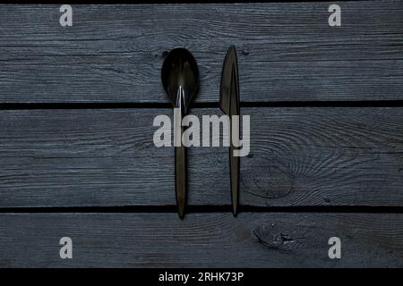 Una cuchara de plástico y un cuchillo yacen en una tabla de madera negra, vajilla desechable Foto de stock