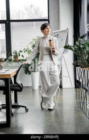 longitud completa de elegante empresaria de mediana edad con café para ir de pie en la oficina moderna Foto de stock