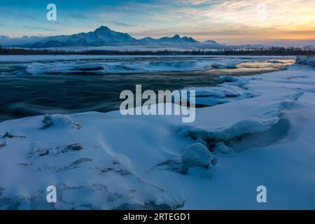 El sol se pone detrás de Pioneer y Twin Peaks y la cordillera Chugach en una noche de invierno, con un suave y sedoso río Matanuska, bordeado por hielo... Foto de stock