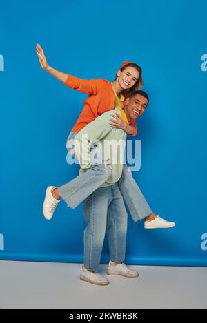 hombre afroamericano feliz piggybacking novia alegre sobre fondo azul, divirtiéndose Foto de stock