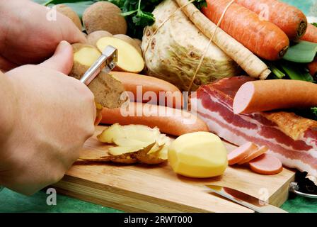 Pele las patatas para la sopa de patatas. Dispuesto delante de verduras frescas, tocino y salchichas Foto de stock