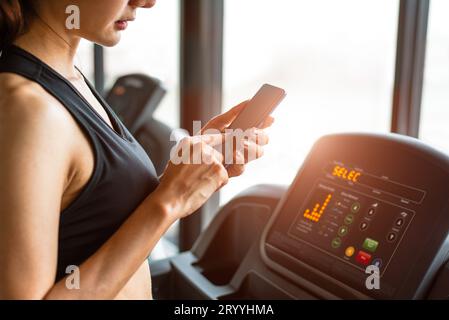Mujer usando teléfono inteligente cuando entrena o entrenamiento de fuerza en gimnasio de fitness en cinta de correr. Relax y concepto de tecnología. Deportes Exer Foto de stock