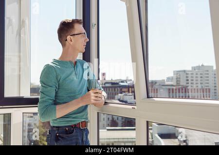 Emociones positivas. Hombre en camiseta turquesa con mangas largas se coloca en el balcón de la casa, apartamento, oficina con vaso de jugo y sonrisas, mirando Foto de stock