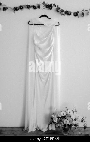 Un elegante vestido de novia blanco exhibido en una pared blanca, adornado con un ramo de flores Foto de stock