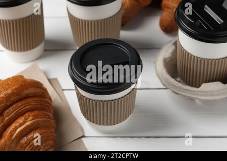 Café para llevar. Tazas de papel con sabrosa bebida y cruasanes en mesa de madera blanca Foto de stock
