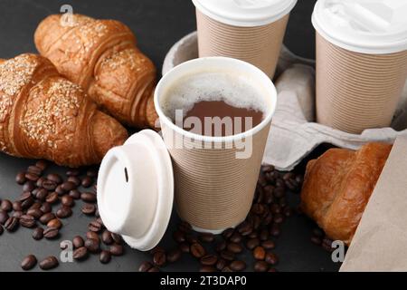 Café para llevar. Tazas de papel de bebida sabrosa, cruasanes y frijoles en mesa negra, primer plano Foto de stock