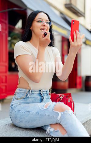 Chica sentada en el banco y mejorando su maquillaje mientras mira en el teléfono inteligente Foto de stock
