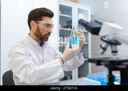 El hombre serio y concentrado maduro trabaja dentro del laboratorio, el científico lleva a cabo experimentos examinando el microscopio. Foto de stock