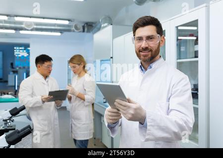 Retrato de técnico de laboratorio experimentado maduro, trabajador de laboratorio sonriendo y mirando a la cámara, hombre en gafas y guantes protectores conduce experimentos, utiliza la computadora de la tableta. Foto de stock