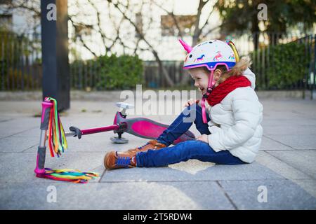 Niña preescolar en casco de unicornio sentado en el suelo después de que se cayó mientras montaba su scooter en el parque en un día de primavera. Actividades deportivas al aire libre y. Foto de stock