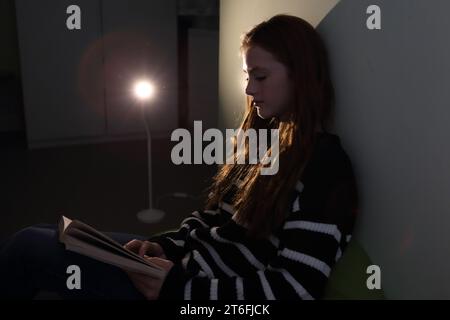 Adolescente leyendo una novela en su dormitorio iluminado por una lámpara Foto de stock