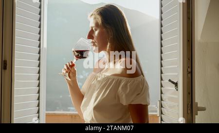 Señora se encuentra en la terraza de la villa, bebiendo vino tinto de una copa, mientras que las montañas escénicas sirven como un telón de fondo cautivador. Foto de stock