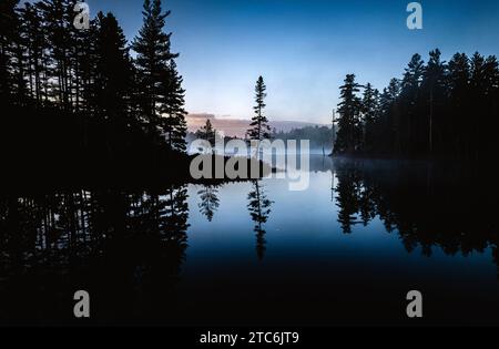 Pinos reflejados en aguas pacíficas del estanque a la hora azul, Maine Foto de stock